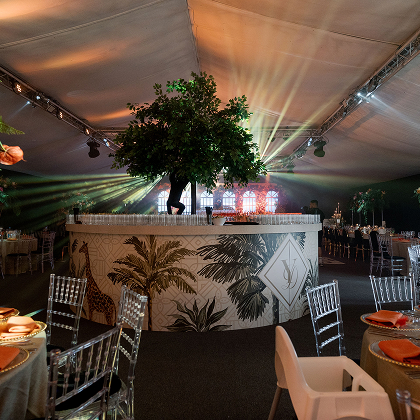 Elegant event setup with tropical-themed bar, greenery centerpiece, and ambient lighting in a decorated venue tent.
