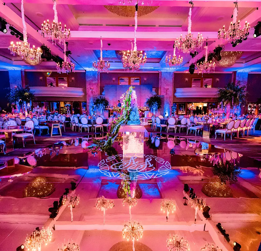 Elegant ballroom with vibrant pink and blue lighting, floral centerpiece, and chandeliers for a lavish event.