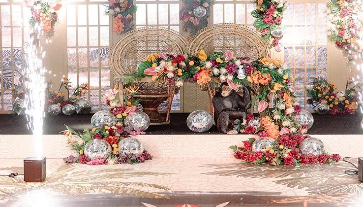 Vibrant decorative stage with floral arrangements, disco balls, wicker chairs, and a monkey statue at a festive event.
